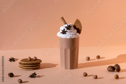 Glass with chocolate cocktail with whipped cream garnished with star anise and cinnamon stick placed on table with hazelnuts and cookies on pastel brown background photo