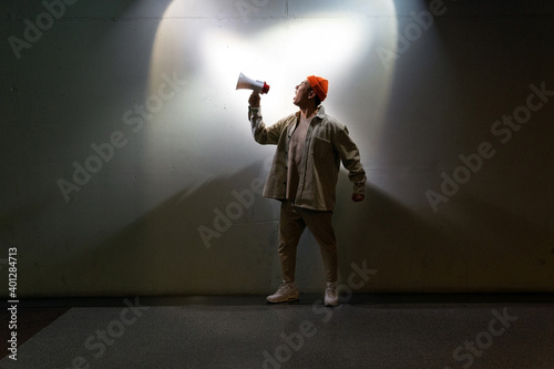 Full body overwhelmed male in casual clothes screaming in loudspeaker while standing against gray wall in dark room photo