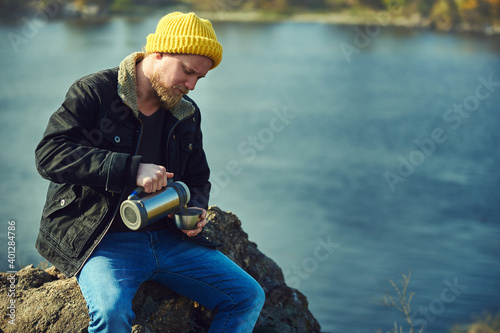 Bearded adventurer man pouring tea or coffee from thermos in a metal cup while resting on a rock on automn nature background. photo