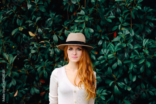 Attractive young long haired ginger female in stylish hat looking at camera while standing against green bushes in summer garden