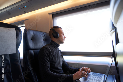 Side view of young male freelancer sitting on passenger seat in modern train and typing on netbook while working during trip