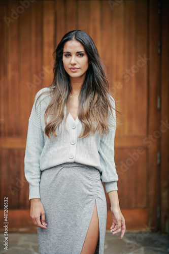 Carefree female with long brown hair and in casual dress standing near wooden doors in city and calmly looking at camera