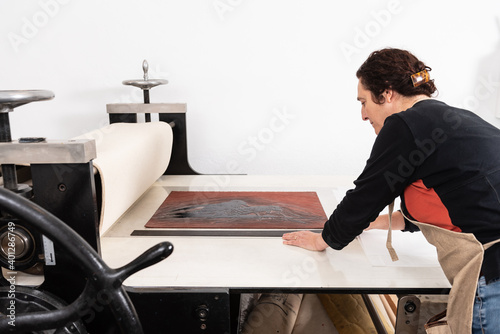 Focused middle aged female master putting sheet of linoleum with inked ornament on printing press while working in linocut technique in work studio photo