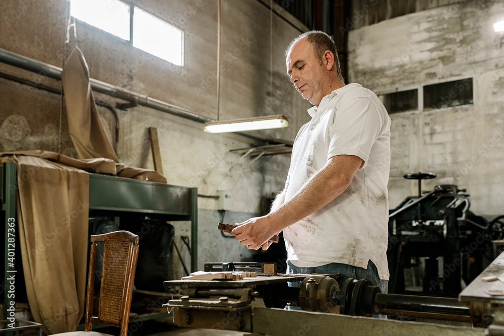 Low angle side view of concentrated male woodworker creating wooden details while standing in grungy garage at daytime Low angle side view of concentrated male woodworker creating wooden details while standing in grungy garage at daytime