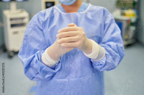 Anonymous female medic in sterile mask and gloves looking at camera in hospital behind medical equipment