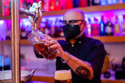 Side view of serious barkeeper in eyeglasses pouring beer into glass while working in bar during coronavirus pandemic photo