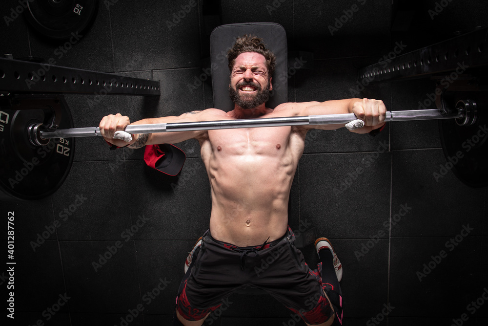 From above of strong male athlete lying on bench press and doing exercises with heavy barbell during workout in gym From above of strong male athlete lying on bench press and doing exercises with heavy barbell during workout in gym