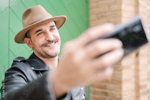 Cheerful glad bearded gallant male in hat taking photo in yard of brick house photo