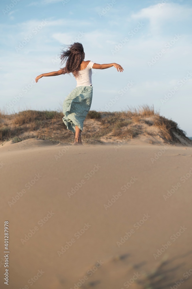 Back view of anonymous graceful ethnic female with curly hair in summer outfit standing on sandy hill with flying hair and outstretched arms while enjoying evening Back view of anonymous graceful ethnic female with curly hair in summer outfit standing on sandy hill with flying hair and outstretched arms while enjoying evening