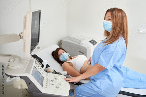 Side view of female doctor in uniform examining patient in mask while watching ultrasound scanner monitor in clinic