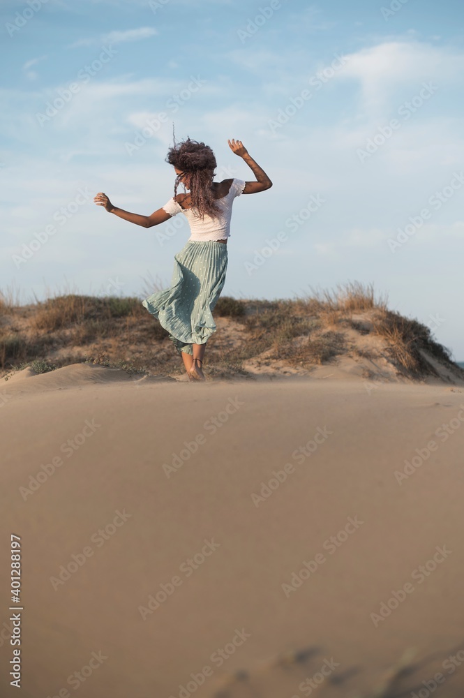 Back view of anonymous graceful ethnic female with curly hair in summer outfit standing on sandy hill with flying hair and outstretched arms while enjoying evening Back view of anonymous graceful ethnic female with curly hair in summer outfit standing on sandy hill with flying hair and outstretched arms while enjoying evening