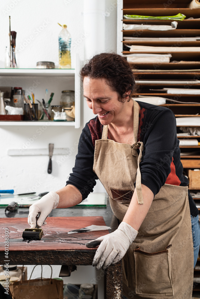 Mature female artist in apron and gloves covering engraved plate with ink while creating artwork in professional workroom Mature female artist in apron and gloves covering engraved plate with ink while creating artwork in professional workroom