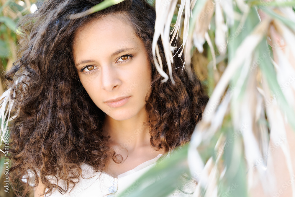 Flirty young ethnic mixed race female with curly hair looking at camera standing in foliage of verdant tree looking at camera Flirty young ethnic mixed race female with curly hair looking at camera standing in foliage of verdant tree looking at camera