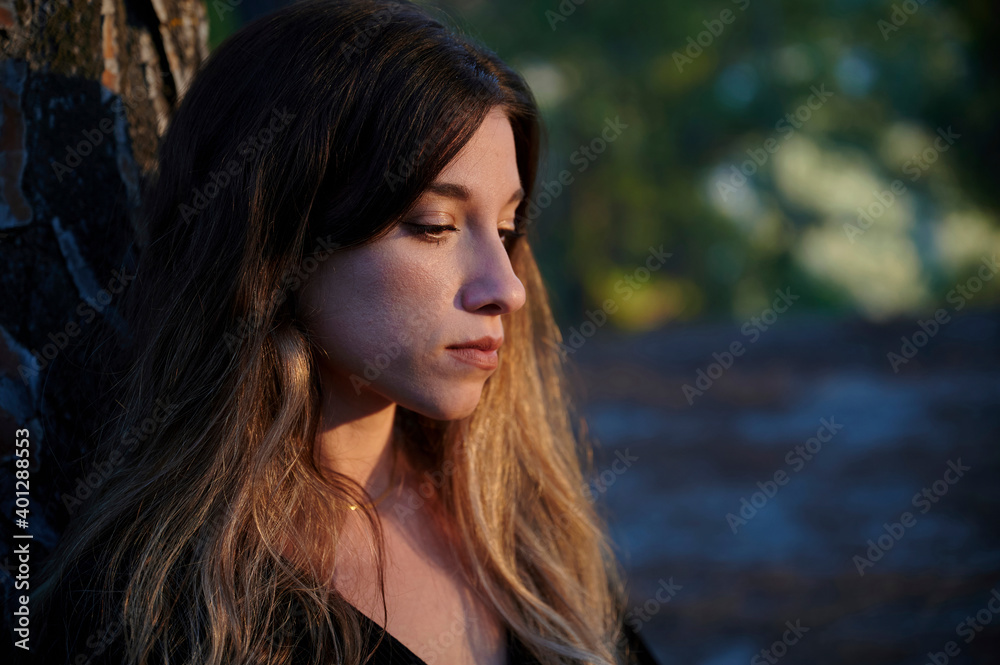 Side view of silent sad young long haired female leaning back on tree trunk while standing against blurred forest Side view of silent sad young long haired female leaning back on tree trunk while standing against blurred forest