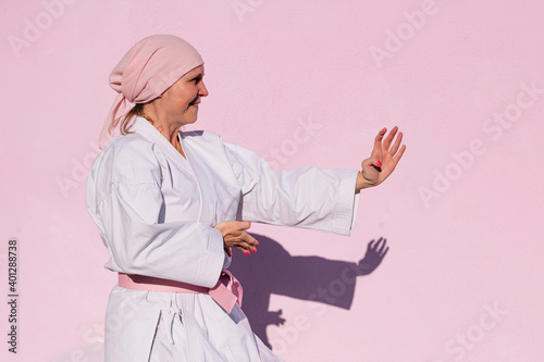 Side view of determined mature woman in pink head cover and belt fighting karate in cancer battle concept in the street on pink wall looking away