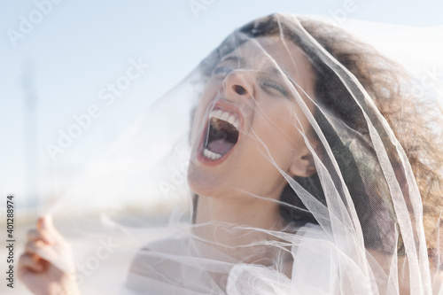Young angry female with curly hair screaming through transparent veil on sunny clear day