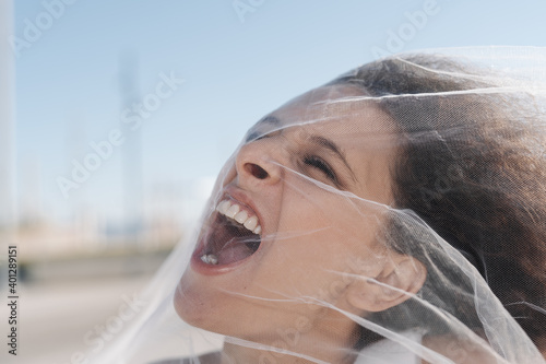 Young angry female with curly hair screaming through transparent veil on sunny clear day