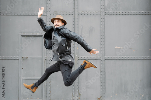 Full body of stylish thoughtful male with mustache in hat leaping and looking at camera near building with metal door and wall
