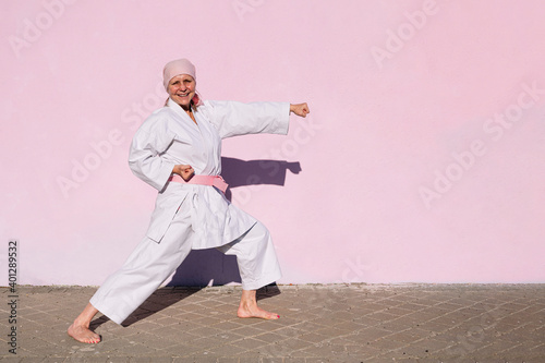 Side view of determined happy mature woman in pink head cover and belt fighting karate in cancer battle concept in the street on pink wall looking at camera photo