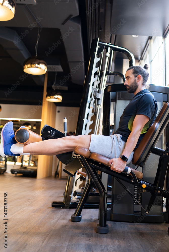 Side view of bearded male athlete doing exercise on leg extension machine during fitness workout in gym