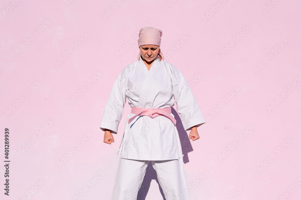 Determined thoughtful mature woman in pink head cover and belt fighting karate in cancer battle concept in the street on pink wall with eyes closed Determined thoughtful mature woman in pink head cover and belt fighting karate in cancer battle concept in the street on pink wall with eyes closed