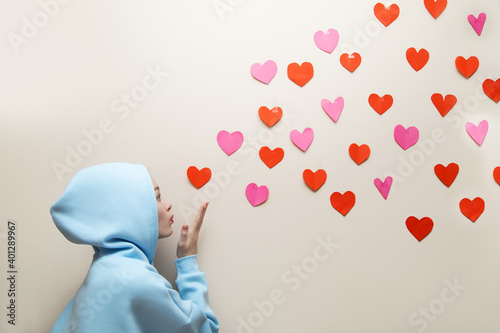 Young Girl in blue hoodie with hood blows off hearts from her palm and they fly along wall, copy space. Valentine's day, search love concept, giving good photo