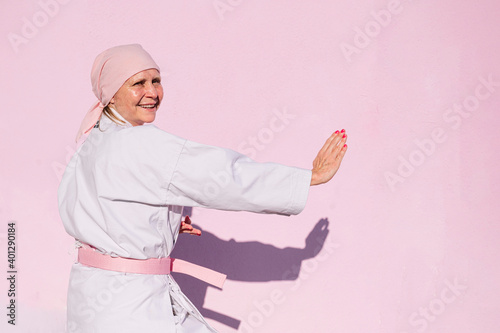 Side view of determined happy mature woman in pink head cover and belt fighting karate in cancer battle concept in the street on pink wall looking at camera