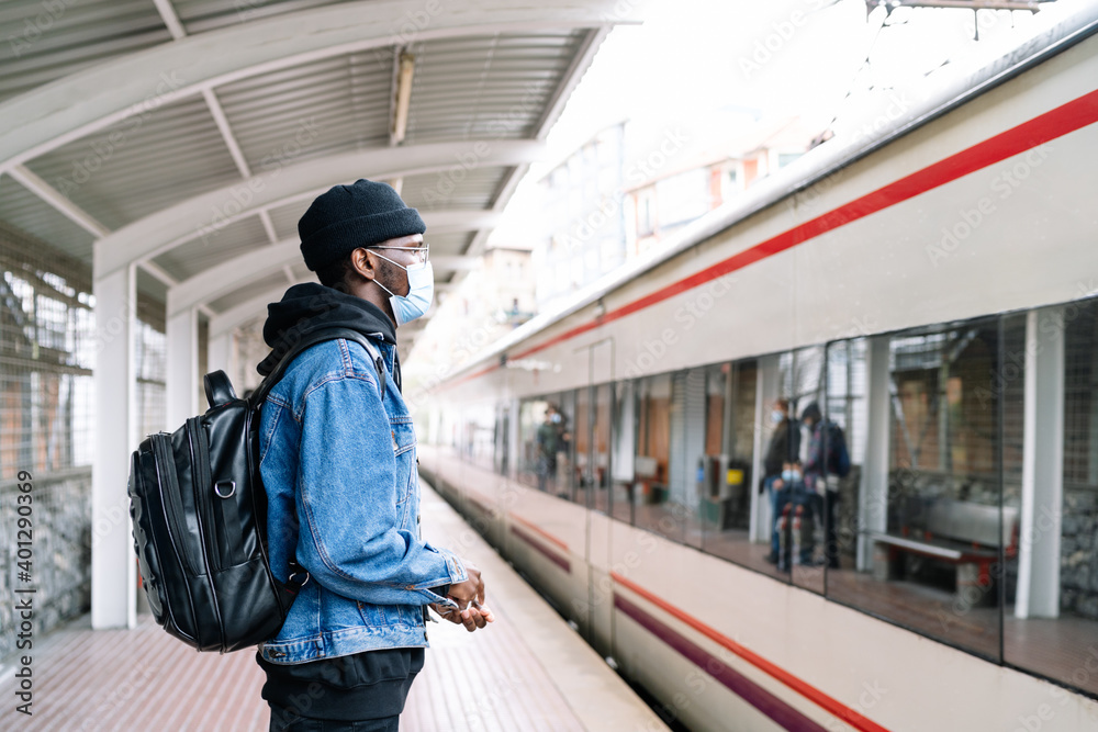 Side view of ethnic male tourist in mask and with backpack standing on platform of railway station during coronavirus epidemic