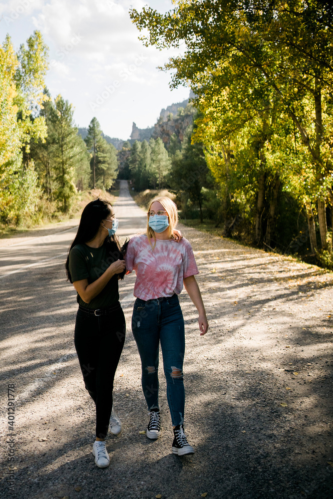 Cheerful young female friends in casual wear and protective masks hugging and having fun while standing on asphalt country road leading through forest in autumn day Cheerful young female friends in casual wear and protective masks hugging and having fun while standing on asphalt country road leading through forest in autumn day