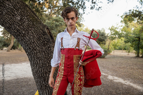 Young matador wearing red traditional costume standing in nature with cape and sword looking at camera photo