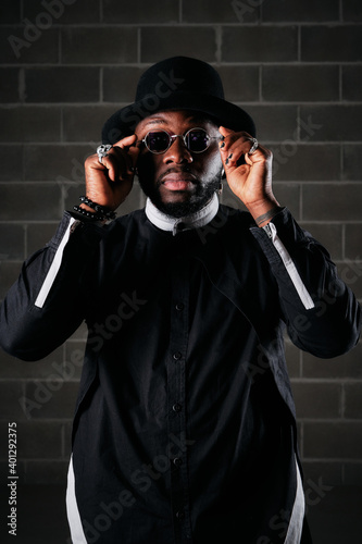 Stylish African American male in black apparel putting on trendy round sunglasses and looking at cmaera while standing on background of brick wall in studio photo