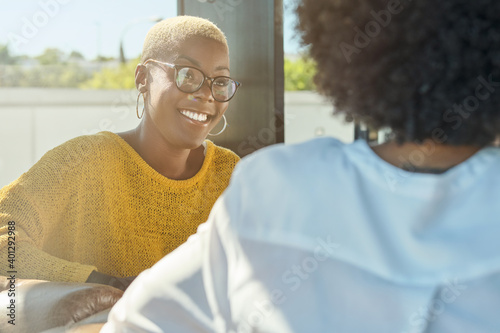 African American female with short hair speaking to unrecognizable friend with curly hair sitting on terrace and talking laughing at jokes and looking at each other while drinking hot beverage