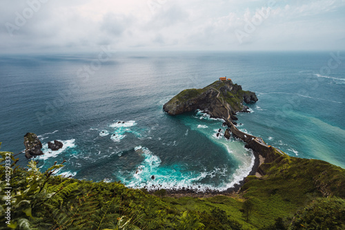Drone view of paving stone way leading along stone bridge and ridge of rocky hill to lonely house on island Gaztelugatxe surrounded by tranquil sea water under cloudy sky in Basque Country photo