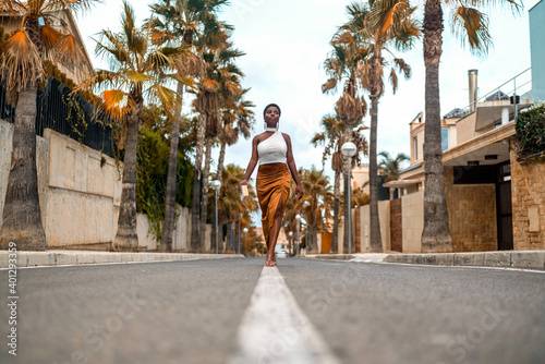 Ground level of young dreamy African American female in trendy apparel strolling on asphalt roadway while looking away photo