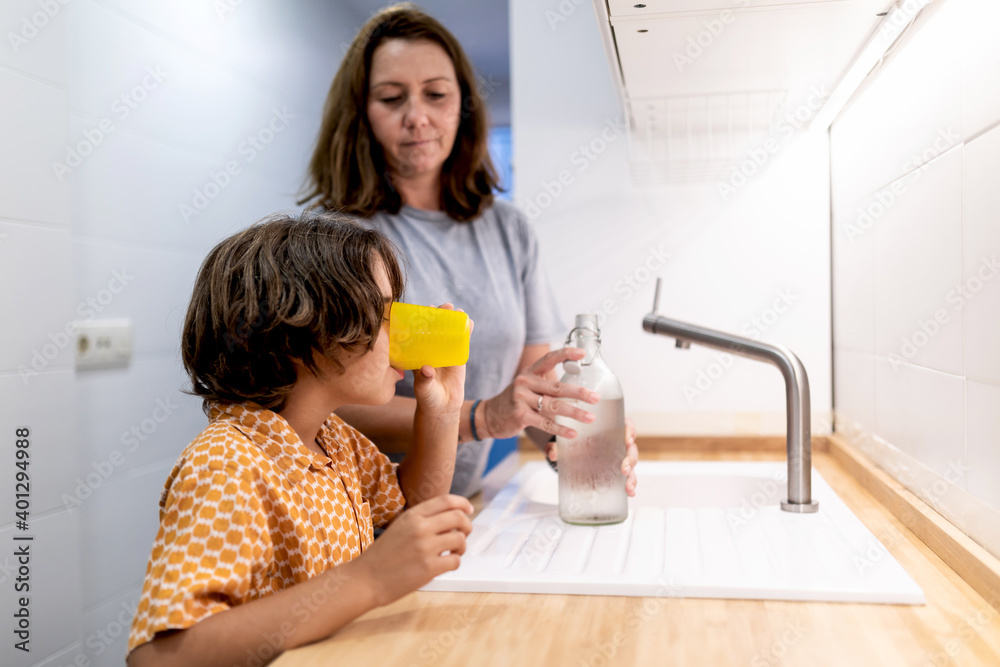 Unrecognizable kid in ornamental clothes drinking water from glass near woman with bottle at home Unrecognizable kid in ornamental clothes drinking water from glass near woman with bottle at home