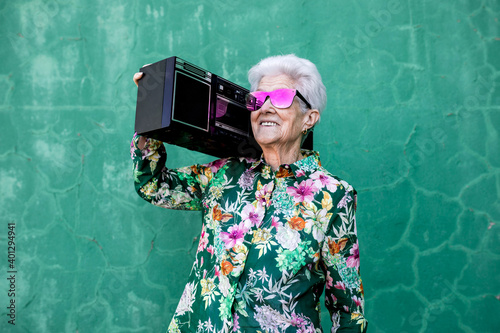 Cool elderly female in colorful trendy blouse and sunglasses carrying record player and enjoying music against green wall photo