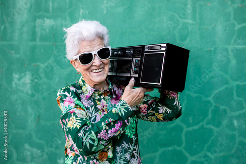 Happy elderly gray haired female in stylish outfit carrying record player on shoulder while standing against green wall on street looking at camera photo