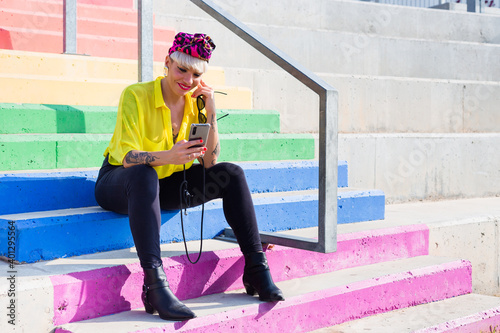 Smiling female in funky outfit sitting on colorful rainbow stairs and browsing smartphone during weekend in city photo