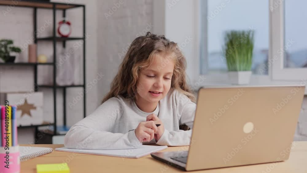 Cute primary school girl studying at home using laptop computer. Schoolgirl has online lesson, writing in exercise book, doing homework. Distance learning online. Children home education. Cute primary school girl studying at home using laptop computer. Schoolgirl has online lesson, writing in exercise book, doing homework. Distance learning online. Children home education.