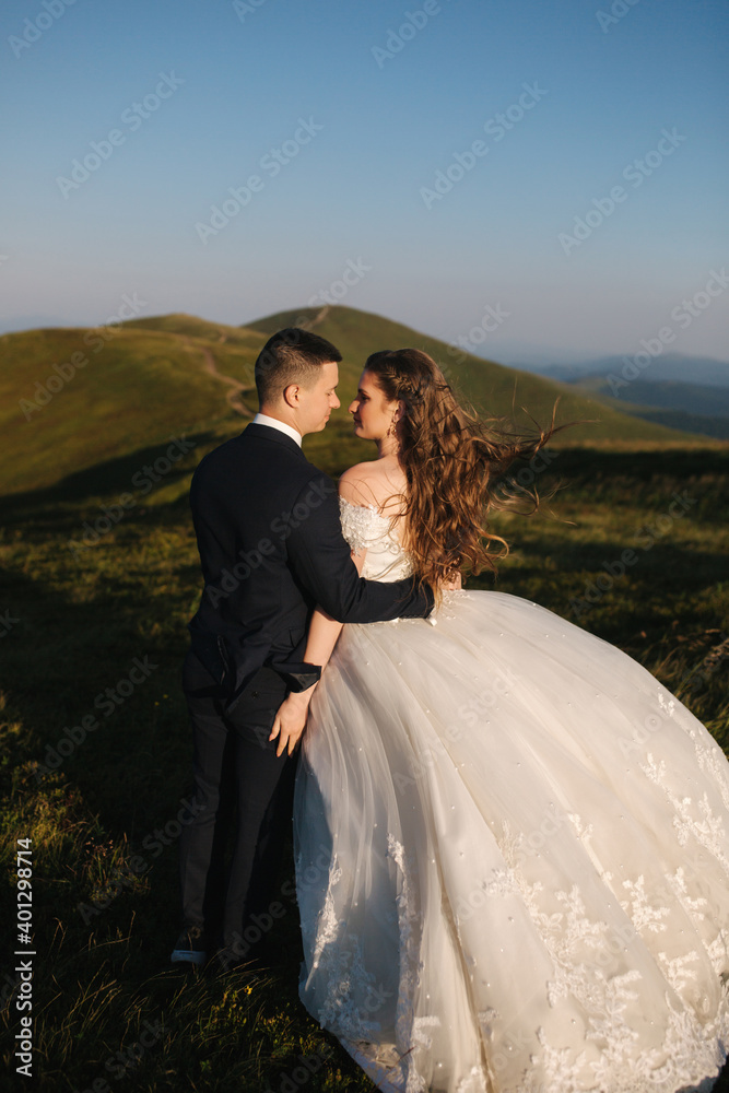Groom and bride on the hill in mountains. Wedding couple in front of green hill. Newlyweds enjoying romantic moments in the mountains at sunset in beautiful summer day