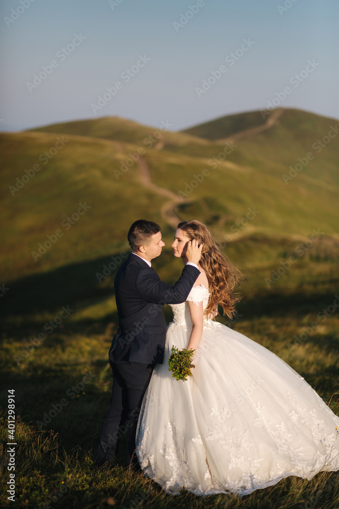 Full length body portrait of attractive bride and stylish groom enjoying romantic moments in the mountains at sunset in beautiful summer day. Wedding couple. Green hills on background
