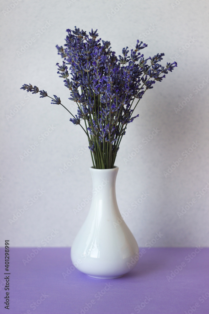 A bouquet of summer lavender flowers stands in a white small vase on a lilac table and against a white wall