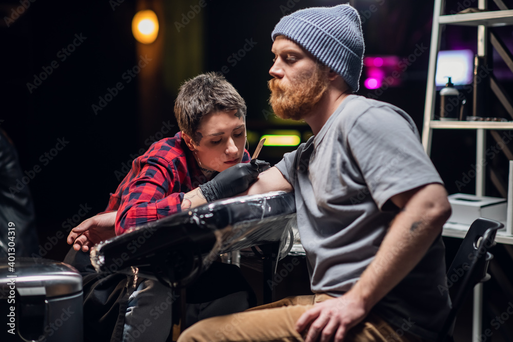 Short-haired girl with piercings and tattoos on her face and hands, the master makes a tattoo in a handpoke way for a young man with a red beard and a knitted hat