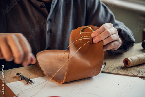 Sitting at a table in the workshop a young guy manually sews leather elements to each other.