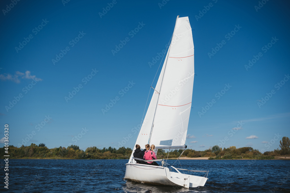 Obraz premium A beautiful white yacht is sailing in the wind on the river against the background of a beautiful autumn forest. A guy on board with two girls