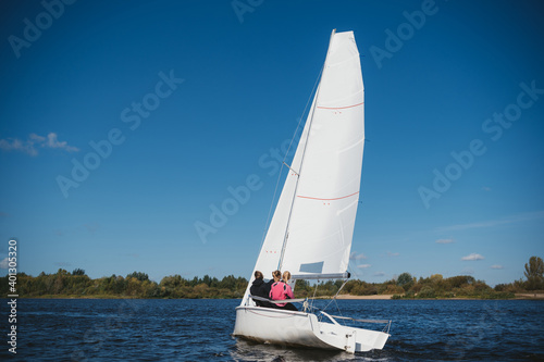 A beautiful white yacht is sailing in the wind on the river against the background of a beautiful autumn forest. A guy on board with two girls © Anna Kosolapova
