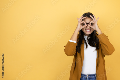 Young beautiful woman wearing a blazer over isolated yellow background doing ok gesture shocked with smiling face, eye looking through fingers