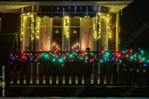 Beautiful multicolored illumination for Christmas and New Year at wooden red Swedish house, window with traditional advent candlestick and wooden terrace fence, snow, foggy night, close view