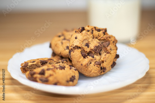 Chocolate chip cookies with a shallow dept of field, with glass of milk in the background, on a wooden table.