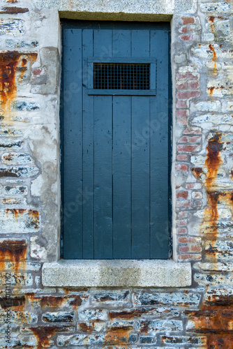 A bright blue vintage exterior wooden door with a small window opening on the top. The entrance is in an old rustic textured rock wall. There are rust stains running down sections of the building.  © Dolores  Harvey